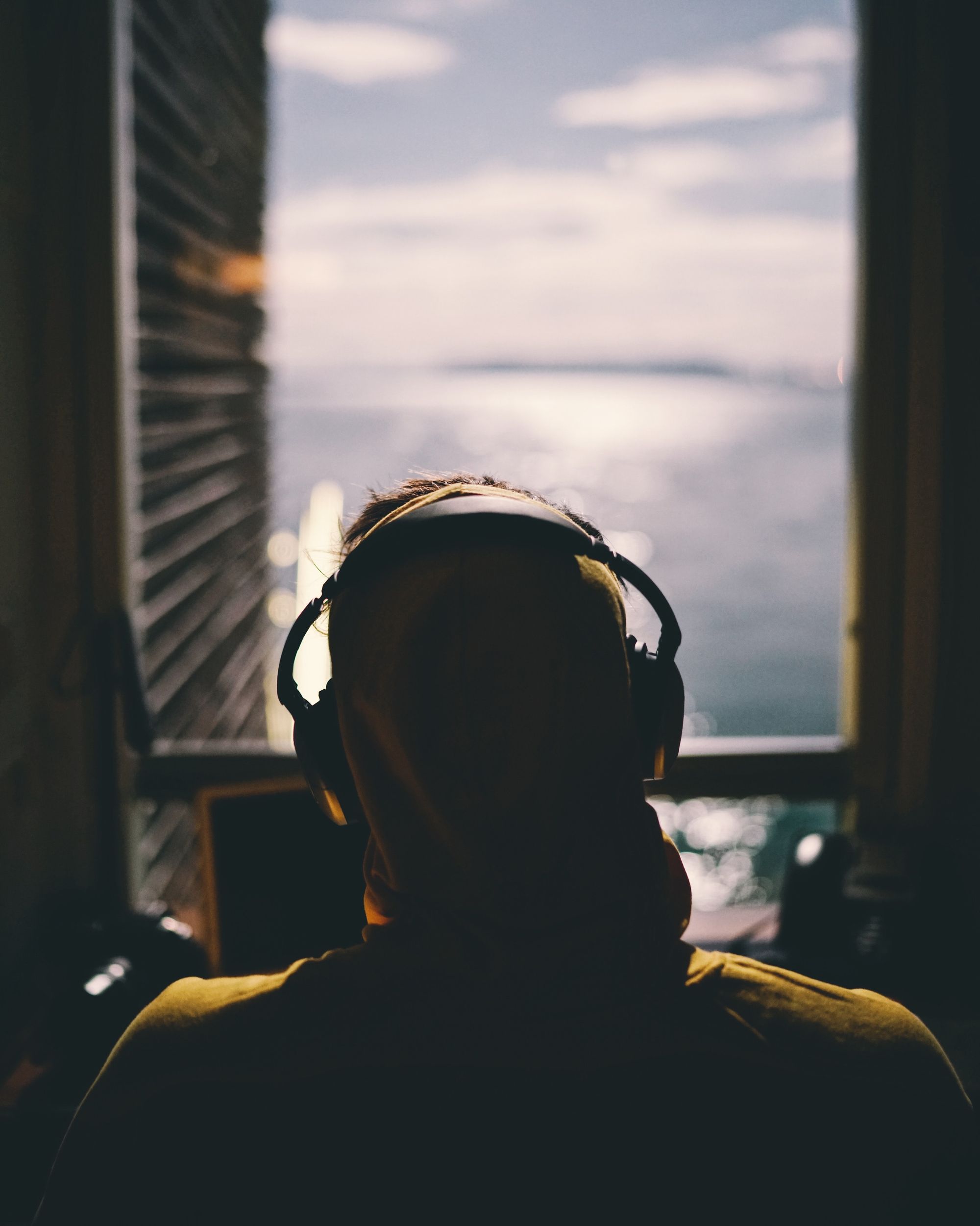 A person wearing headphones sitting down and facing a window, looking out over a body of water.