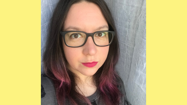 Sarah Frisk headshot, wearing red lipstick and glasses with dark hair dyed pink at the tips.