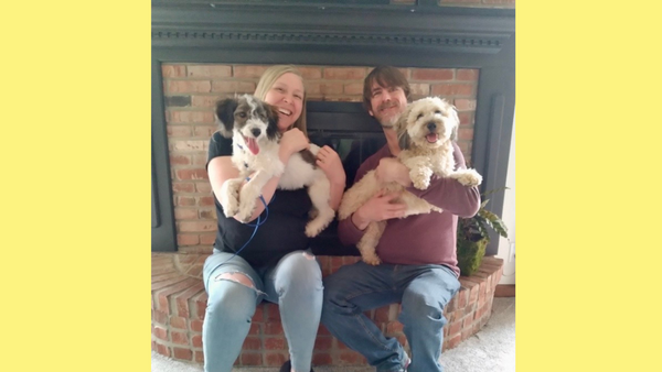 Ann McHealy sitting in front of a fireplace with her husband. Each of them is holding a fluffy dog.