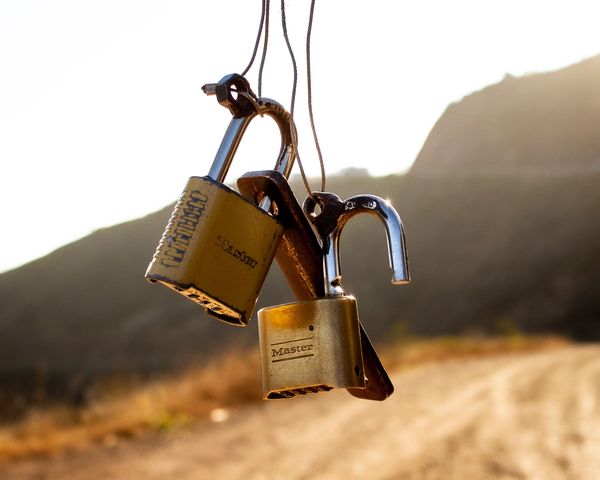 Two brass padlocks hanging from an old latch and wire. One lock is open. There are mountains in the background.
