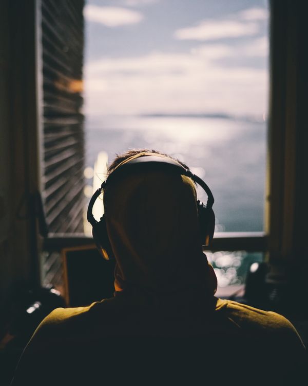 A person wearing headphones sitting down and facing a window, looking out over a body of water.