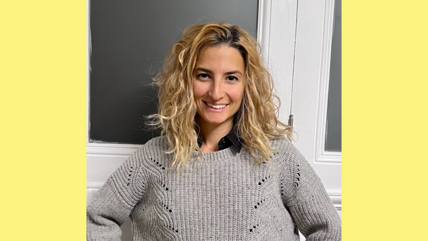 Zeynep, a smiling blonde woman with riotous mermaid curls, in a paneled grey and white room and cozy sweater.